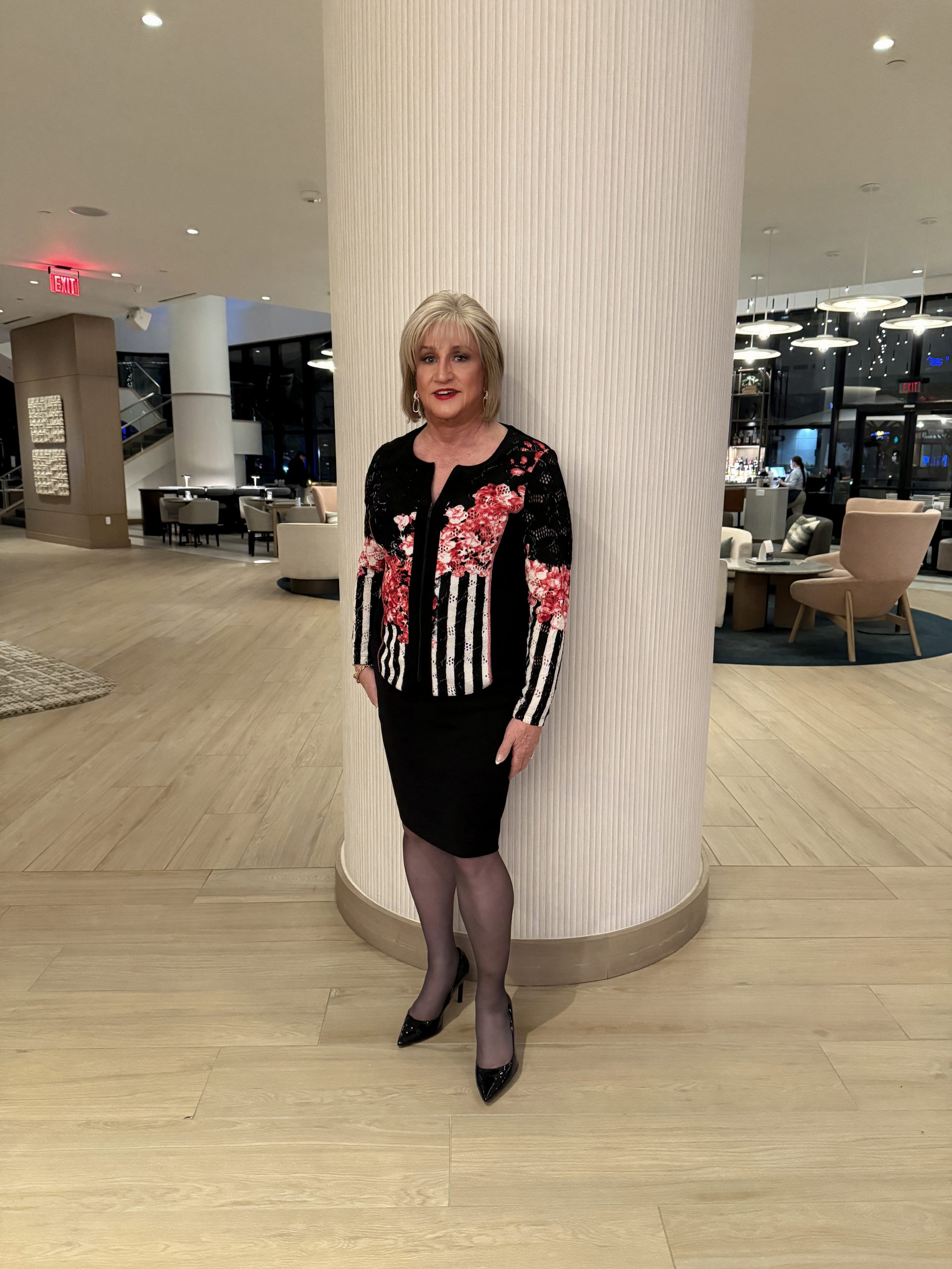 A woman standing in a modern interior space, wearing a floral patterned black and white sweater with a black skirt and high heels, leaning against a textured column.